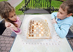 Little frowning girl and smiling boy play chess at table in park