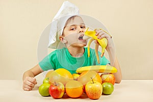 Little funny boy eat fresh banana at table with fruits