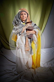 Little funny boy in long white and yellow dress with scarf on head and jug. Young male model posing for photo shoot in