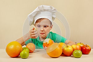 Little funny chef peeling fresh orange at table with fruits