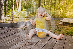 Little funny cute blonde girl child toddler with dirty clothes and face eating baby food fruit or vegetable puree with