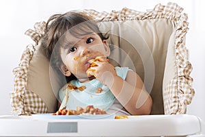 Little funny girl of 12 months eating spaghetti with spoon while sitting in high-powered chair at home. Toddler child