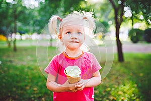 Little funny girl blonde eating sweet blue ice cream in a waffle cup on a green summer background in the park. smeared her face an