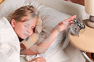 Little funny girl lying in bed with hand turns off the alarm clock waking up in the morning.