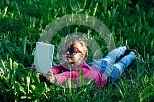 Little funny girl makes a selfie on a tablet in the green grass