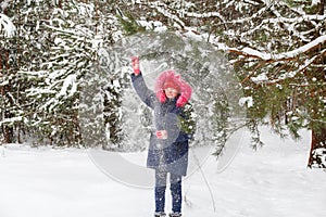 Little funny girl in red warm hat shakes the snow from a branch outside on nature winter snowy forest background. Pretty