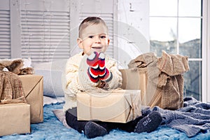 Little funny playful boy a child sits on a bed on Christmas day with gift boxes in white wool knitted sweater and big bright mitte