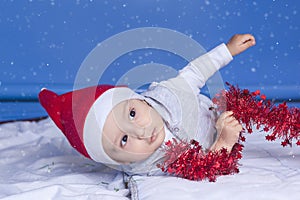 Little funny Santa boy playing with christmas or new year holiday festive garland. 1 year old baby boy in Santa Claus cap