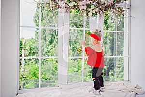 Little funny two years old baby girl in stylish red clothes and jeans and sneakers stands with back on the windowsill near the win