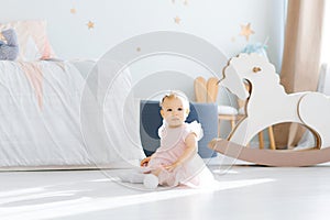 A little girl 1 year old in a pink dress sits on the floor in the children`s room