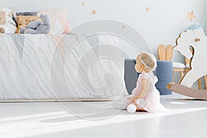 A little girl 1 year old in a pink dress sits on the floor in the children`s room and looks at the bed