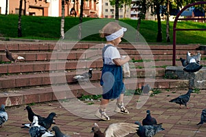 Little girl, 18 months old, in a white T-shirt, denim overalls, white rim running near pigeons, chasing pigeons. Baby feeds