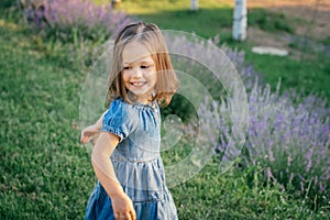 Little girl 3-4 with dark hair in denim dress in sun dancing or running and smiling among large bushes of lilac lavender