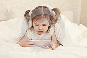 Little girl 3 years old lies under the covers in bed cheerful with smartphone in her hands on white bed, lifestyle