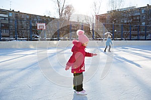 Little girl of 5 years old is skating on ice skates on a skating rink in the courtyard of the house. Frosty winter sunny day,