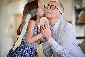 A little girl with angel wings is kissing and exchanging emotions with her grandmother at home. Family, home, love, playtime