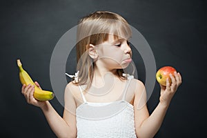 Little girl with apple and banan in the hands. The choice between fruits. Facial expression photo