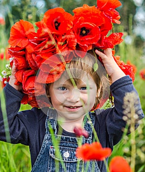 Little girl baby playing happy on the poppy field with a wreath, a bouquet of color A red poppies and white daisies, wearing a den