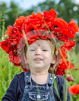 Little girl baby playing happy on the poppy field with a wreath, a bouquet of color A red poppies and white daisies, wearing a den