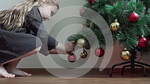 A little girl with bare feet coming to the Christmas tree and hang a garland on a branch in the form of New Year`s decorations