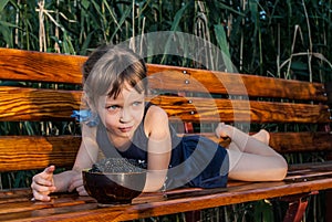 A little girl with beautiful big blue eyes lies on the bench with a bowl of fresh blachberries in front of her.