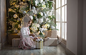 A little girl in a beautiful dress is sitting under a Christmas tree with a gift box and a bow. The light from the large window, t