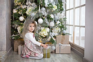 A little girl in a beautiful dress is sitting under a Christmas tree with a gift box and a bow. The light from the large window, t