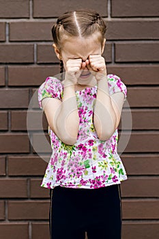 A little girl with a beautiful hairstyle is very upset and cries, wiping her eyes with her hands