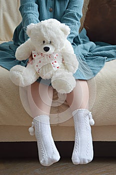 A little girl in a turquoise dress holds a white toy bear with a ribbon with hearts in her hands. a child`s toy.