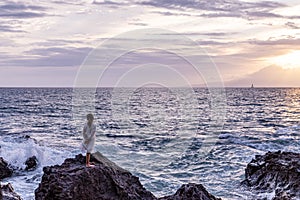 A little girl in a beautiful white dress standing on a rock in front of the ocean watching sunset