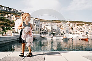 Little girl with a big pink plush hare stands on the pier against the background of the marina with yachts