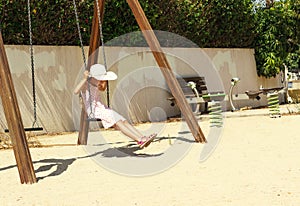 Little girl in a big white hat having fun on a swing at sunny day