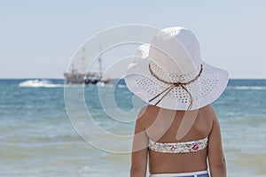 Little girl in a bikini and a white beach hat looking at the sea