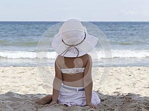 Little girl in a bikini and a white beach hat looking at the sea