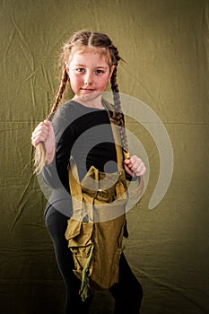 A little girl in a black leotard and a military bag over her shoulder stands against a green background. A child dressed in