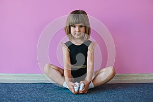 Little girl in a black leotard sitting in a lotus position on a background of a pink wall. Gymnast doll