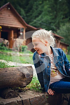 A little girl with blond hair, in a denim jacket, sits on a lawn near a mountain forest, laughs