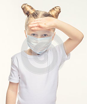 A little girl with blond hair in a medical mask touches her forehead with her hand. Medical concept of epidemiological period,