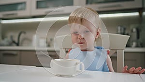 Little girl with blond hair sitting at dining table in kitchen eating homemade food with a spoon.