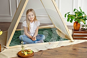 Little girl with blond hair in a white shirt and blue jeans is smiling sitting in a tent in the children`s room.
