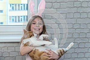 A little girl with blonde hair holds a red cat in her hands