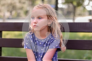 Little girl, blonde, sits with a displeased look, angry, on a bench