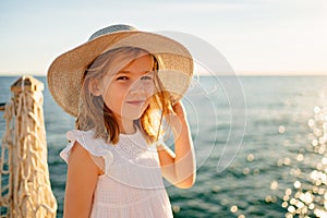 a little girl blonde in a white dress and a wide-brimmed hat stands by the sea