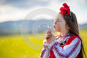 Little girl blowing dandelion on a green field. Bulgarian woman in ethnic folklore dress blow dandelions