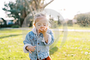 Little girl blows dandelion fluff from her palm while standing in a sunny meadow