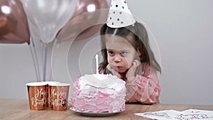 Little girl blows out candle on a birthday cake at home against a backdrop of balloons. Child's birthday.