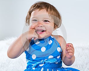 Little girl in a blue dress is sitting and laughing holding her finger in her mouth
