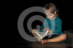 a little girl in a blue dress 4 years old is sitting on the floor in a dark room and reading a book. home schooling in quarantine
