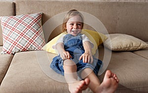 a little girl in a blue jumpsuit lies at home on the sofa among the colorful pillows and smiles cheerfully