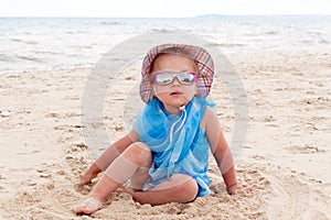 A little girl in blue pareo, sunglasses and a hat sits on the golden sand on the beach by the sea. Tanned skin with